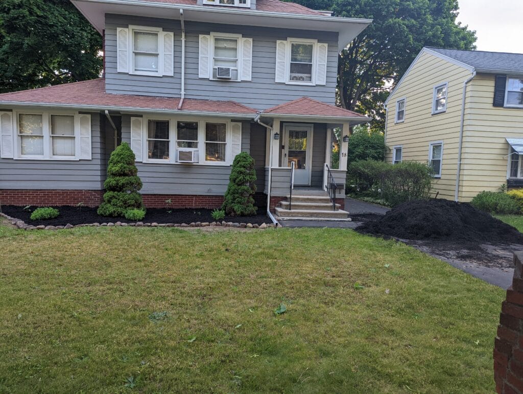 Pile of mulch in driveway and garden beds covered with fresh mulch, showcasing professional mulching services by EJ Lawn & Landscape in Irondequoit, NY